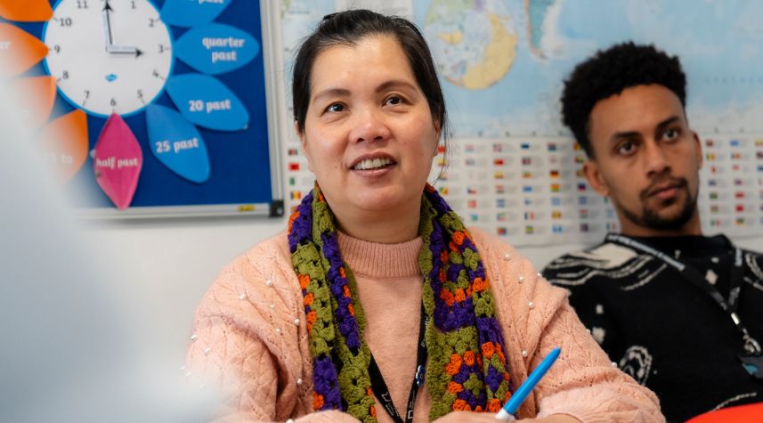 Adult learner seated in a classroom at The Manchester College, listening and writing notes during a lesson, with learning materials displayed in the background.