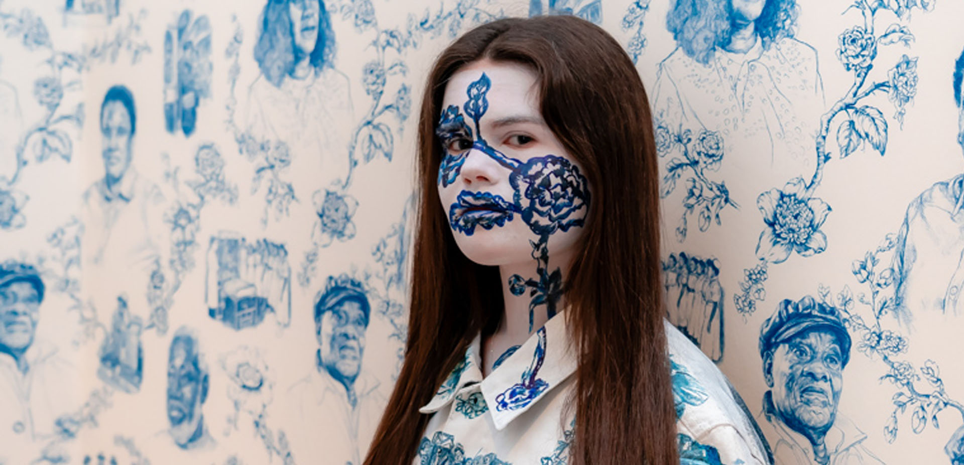 A striking portrait image of a learner with elaborate face paint standing against a wall painted in a similar design