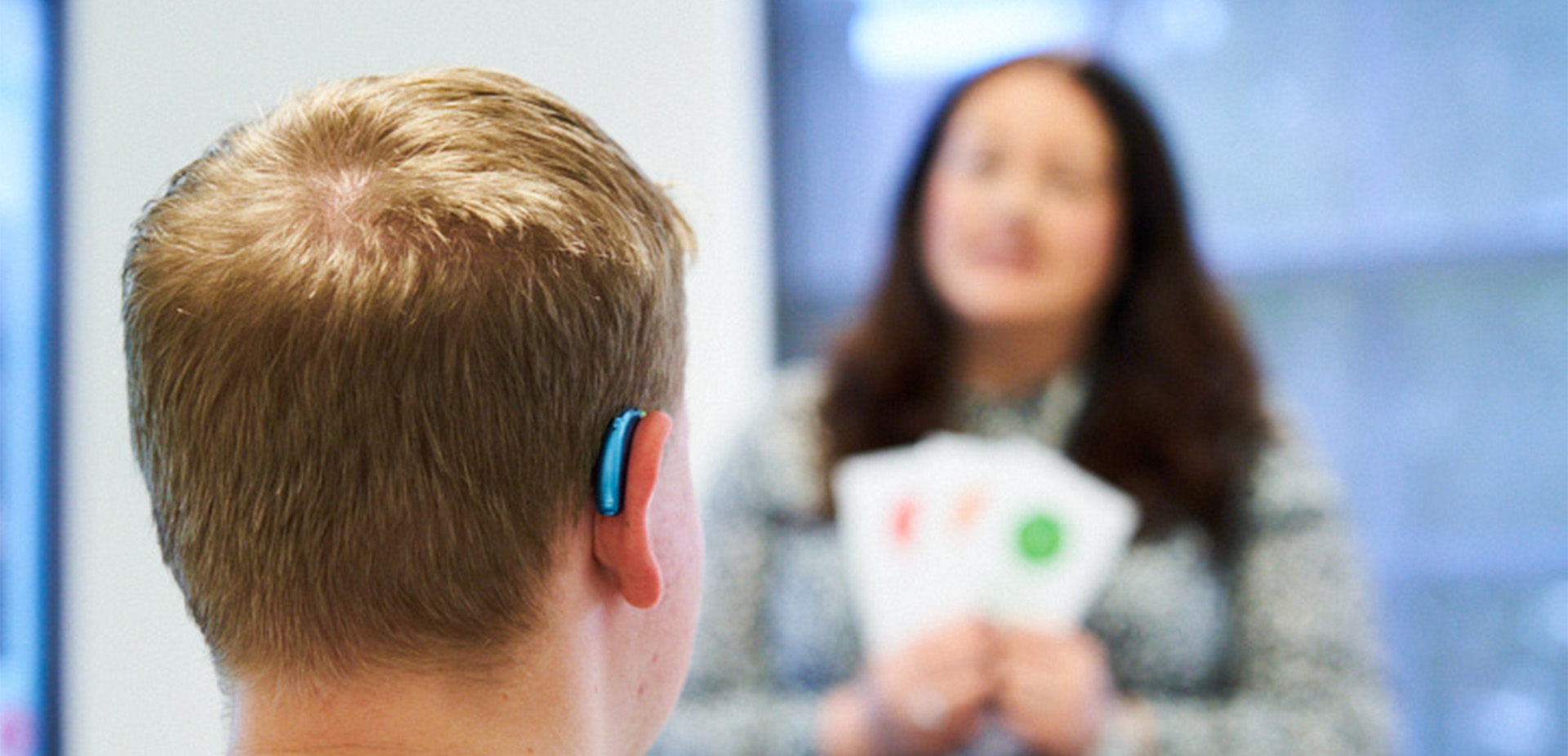 Supported learning session showing a person wearing a hearing aid listening to another person holding coloured communication cards in a classroom setting.