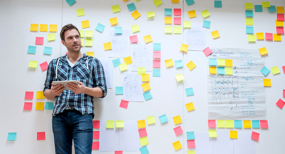 Man standing next to a white board with different coloured post it notes on