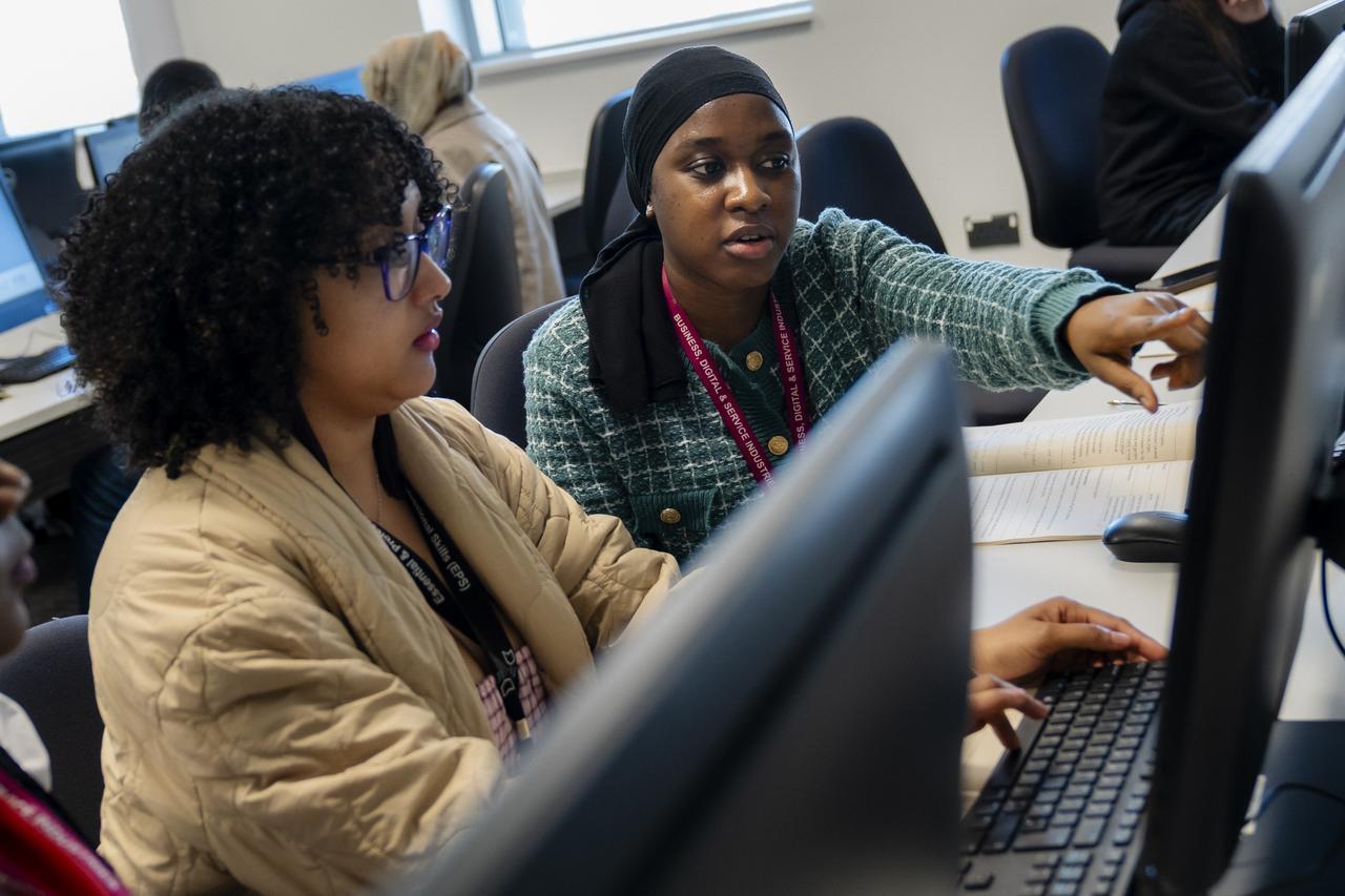 Two TMC students work together at a computer workstation in a classroom, with one student pointing at the screen while discussing work and the other using the keyboard.