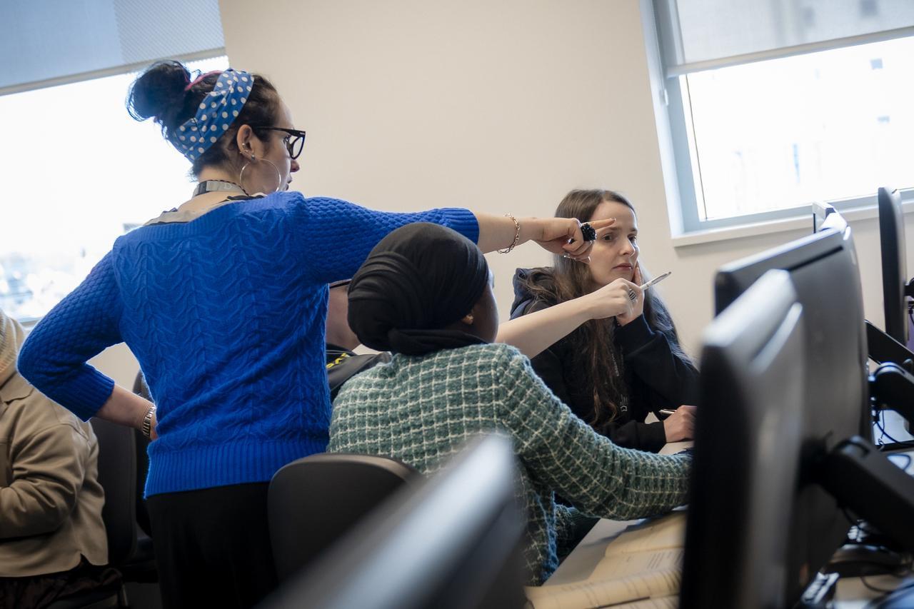 A TMC tutor supports students during a classroom activity, pointing towards a computer screen while students work collaboratively at their workstations.