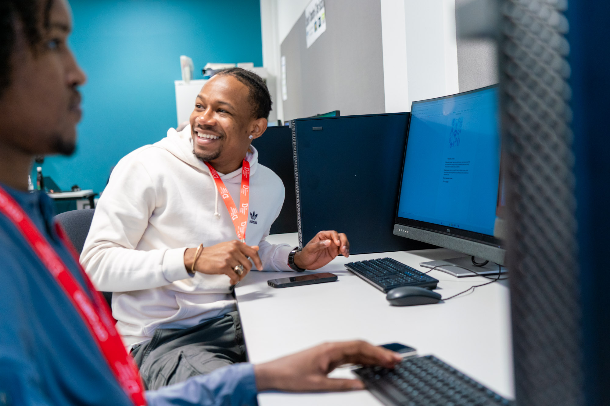 Two students seated at computer workstations in a modern classroom, discussing something on a desktop screen while working with keyboards and other equipment.