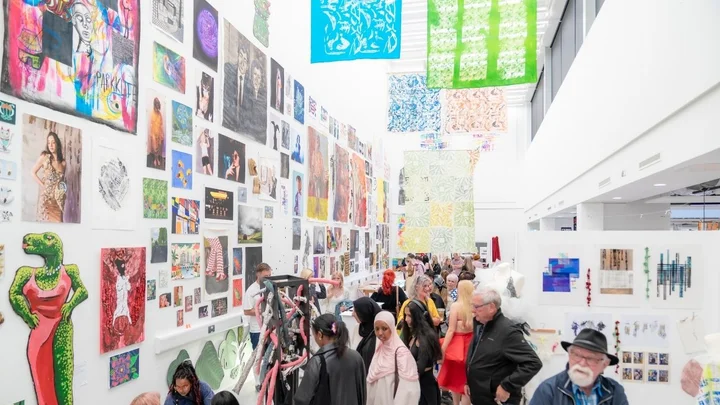 An art exhibition filled with colourful student work displayed on tall gallery walls, with visitors exploring the busy exhibition space.