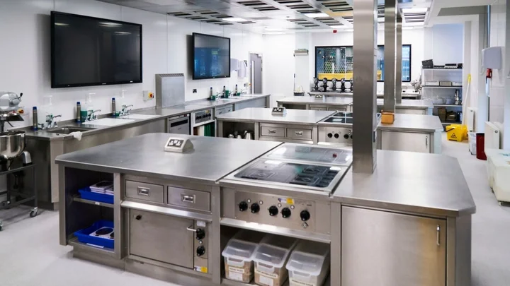 A professional training kitchen with stainless steel workstations, induction hobs, sinks, and mounted screens for teaching demonstrations.