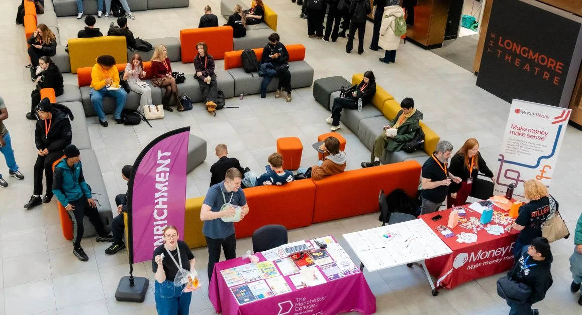 A busy college common area at The Manchester College with students sitting on colourful seating and staff stood at information stalls for enrichment activities.