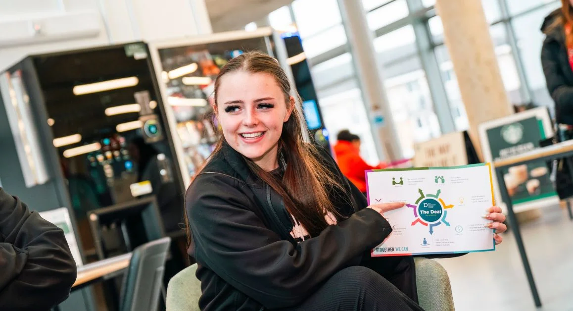 A person seated in in The Manchester College common area holding up a colourful printed sheet titled “The Deal"