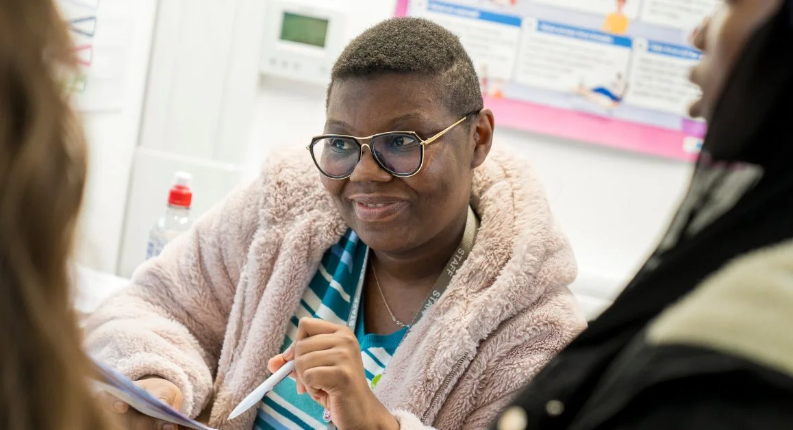 A person sits talking to students, pointing to a sheet of paper, with a colourful wall display visible in the background.