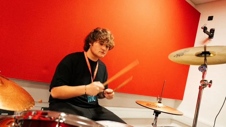 A music learner playing the drum kit during a practice session in a rehearsal room with red acoustic panels.