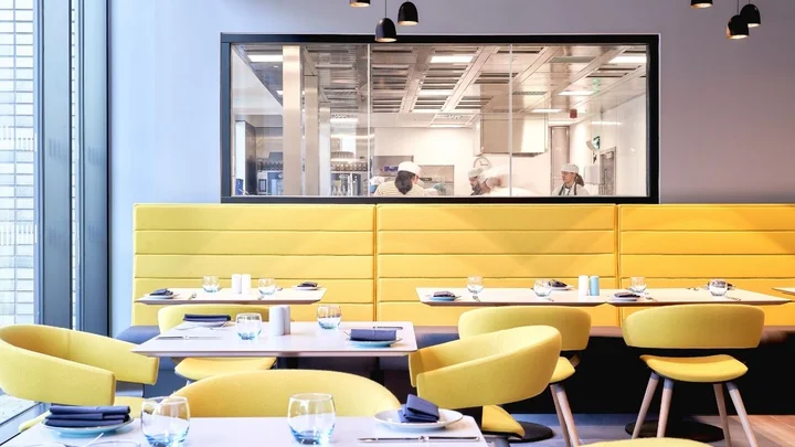 A modern training restaurant with yellow seating and a window overlooking a professional kitchen where catering students are preparing food.