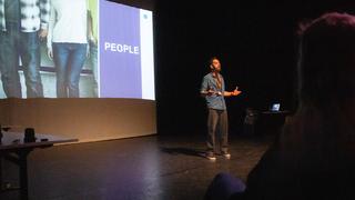 Gurmit Samra stands on stage in a small theatre. He's in front of a projector screen and addressing the crowd.