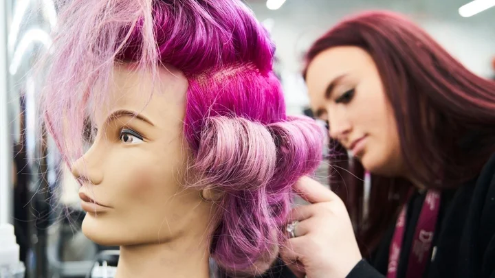 A hairdressing learner styling vibrant pink and purple hair during a practical salon training session.
