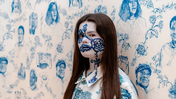 A visual arts learner standing against a hand‑illustrated patterned wall featuring detailed blue line drawings as part of a gallery installation.