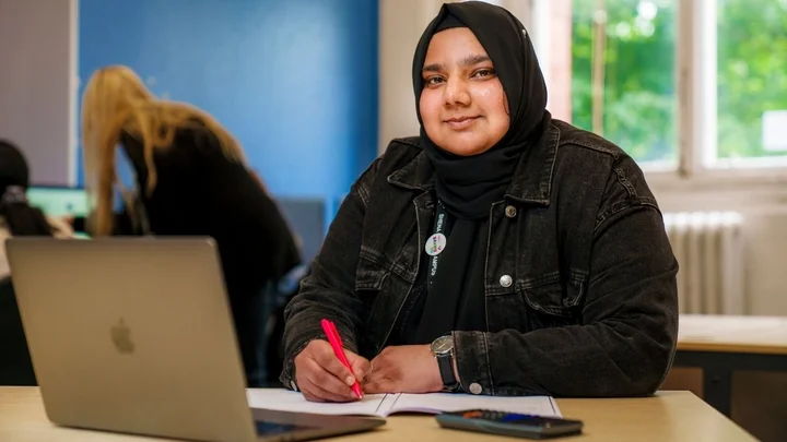 Person seated at a desk using a laptop and writing notes during a business learning session in a classroom environment.