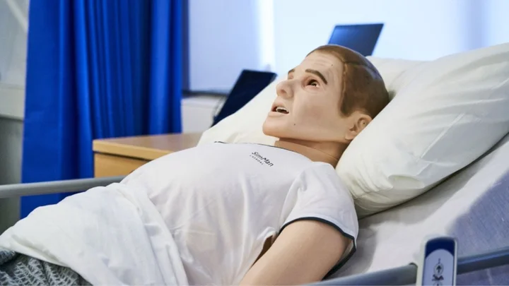 Patient manikin lying in a hospital bed during a clinical skills training session, with medical equipment positioned nearby.