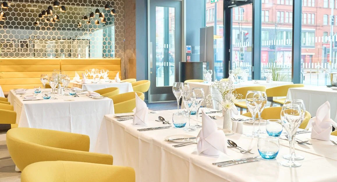 A bright, modern training restaurant at The Manchester College with neatly laid tables, yellow chairs, folded napkins, glassware and cutlery, set beside large windows.