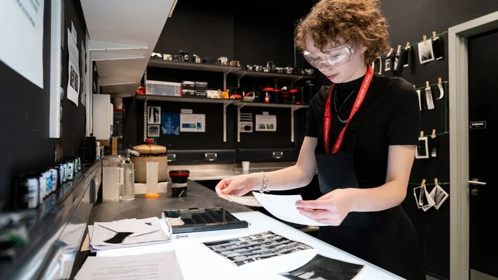 A learner working in a darkroom‑style photography workspace, examining film negatives and prints on a light table with photo‑development equipment nearby.