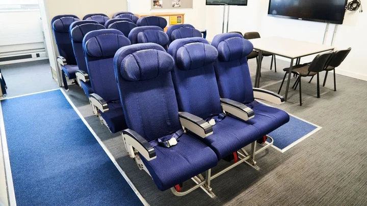 Cabin crew training room featuring rows of blue aircraft seats and a classroom area with tables and chairs.