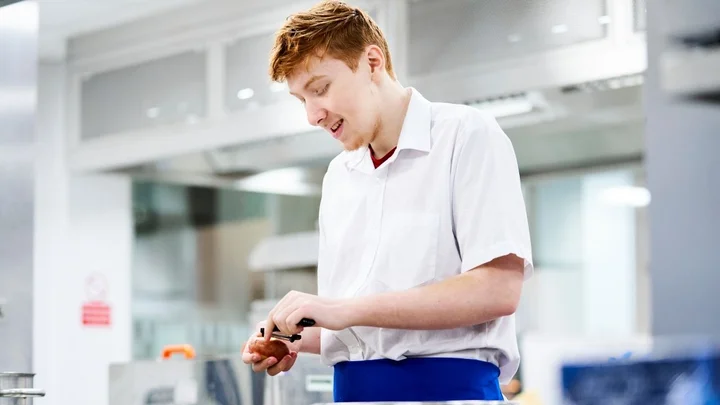A catering learner preparing food in a professional training kitchen, using a peeler at a stainless steel workstation.