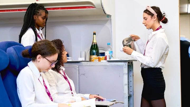 Cabin crew trainees practising in‑flight service, with one learner pouring a hot drink for seated passengers during a simulated cabin environment.
