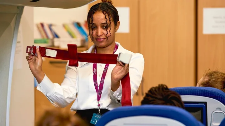 A cabin crew trainee demonstrating a seatbelt safety procedure during a simulated in‑flight training session.