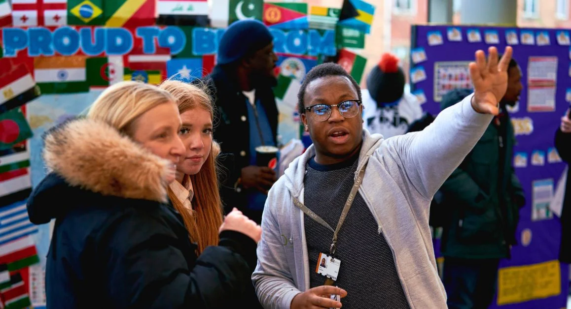 A teacher, student, and parent standing together at a college event, with the teacher gesturing towards a display wall full of information.