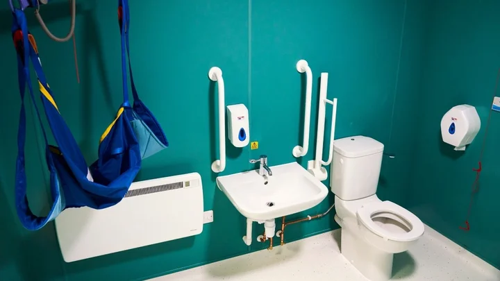 An accessible bathroom with a hoist sling, support rails, sink, toilet, and wall‑mounted dispensers designed for health and social care training.