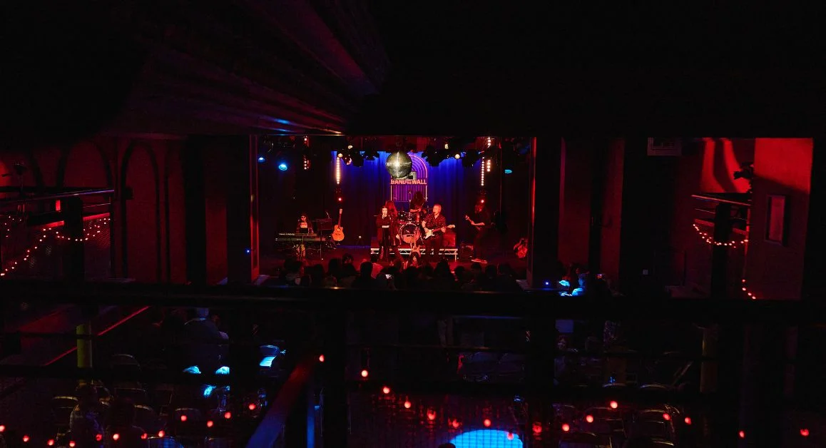 A live music performance on a dimly lit stage viewed from the back of a venue, with musicians playing under red and blue lighting.