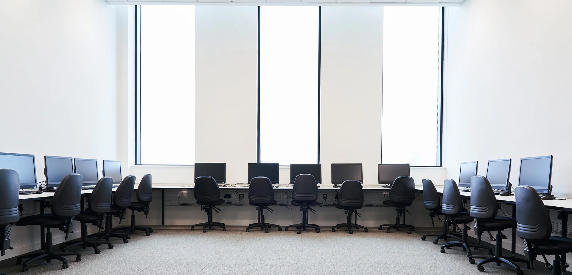 IT training room with rows of desktop computer workstations and chairs arranged around the walls for classroom‑based learning.