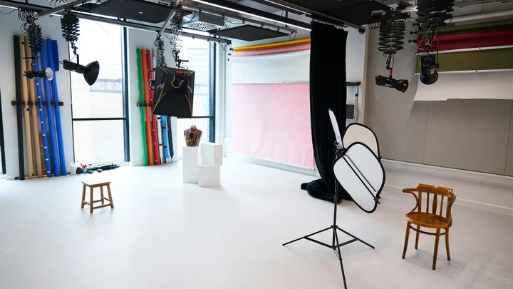 A photography studio setup with softbox lighting, coloured backdrop rolls, reflectors, stools, and display plinths arranged for a photoshoot.