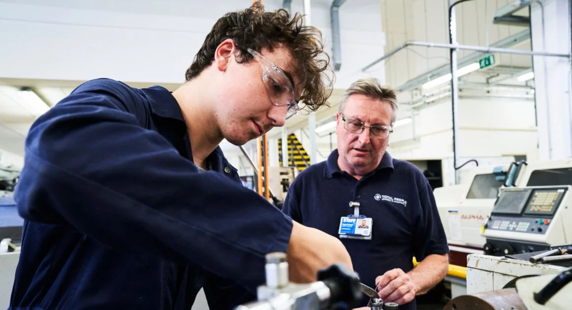 An engineering apprentice receiving hands‑on guidance in a workshop, working with specialist equipment and machinery.