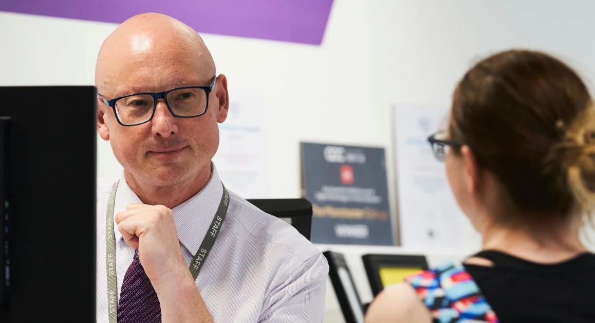 A staff member sitting at a desk in a college support area, speaking with a learner who is seated opposite. Posters and screens are visible in the background.