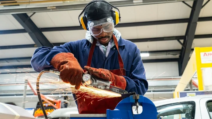 utomotive learner using a grinding tool on a metal component while wearing protective equipment.