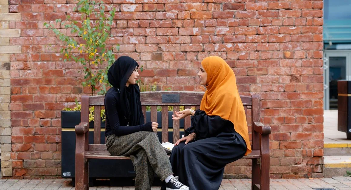 Two individuals sitting on a wooden bench outside a brick building at The Manchester College campus, engaged in conversation.