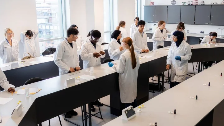Science learners in lab coats working across a laboratory, discussing experiments and preparing equipment at long bench tables.