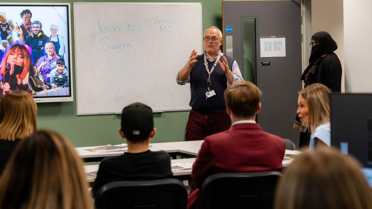 Students attend a classroom session while a tutor presents at the front of the room beside a whiteboard during a college information event.