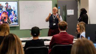 Students attend a classroom session while a tutor presents at the front of the room beside a whiteboard during a college information event.