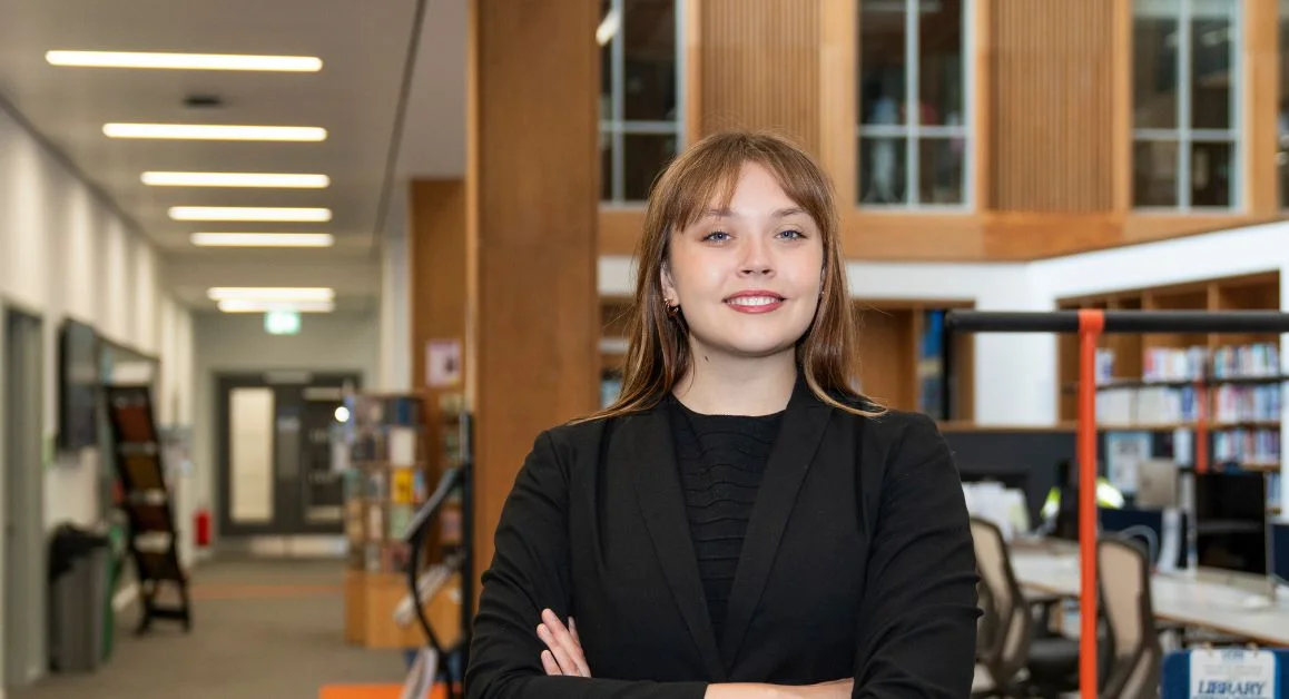 A person standing and smiling in a library with arms crossed, surrounded by bookshelves and desks.