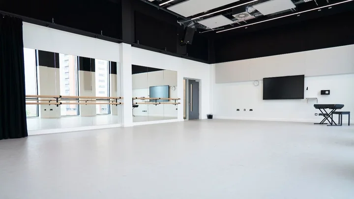A bright dance studio with mirrored walls, ballet barres, a wall‑mounted screen, and a keyboard set up at the side.