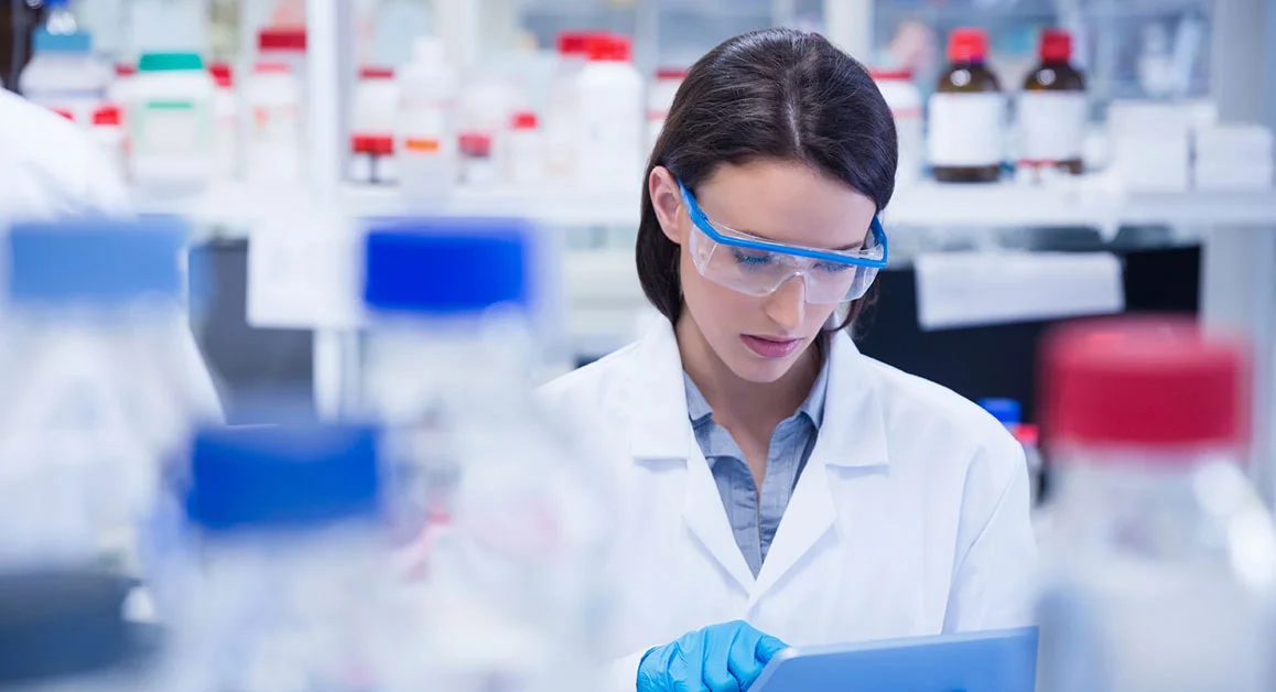 Laboratory researcher wearing safety goggles and gloves working with a tablet in a laboratory setting.