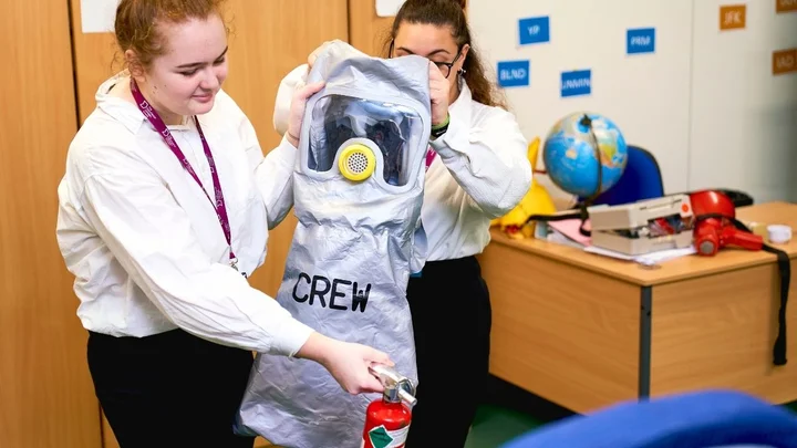 Cabin crew trainees practising emergency equipment handling, demonstrating a protective hood and fire extinguisher during aviation safety training.