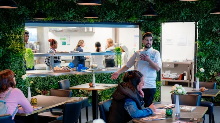 A training restaurant with learners serving customers, while catering students work in the kitchen visible through the pass window.
