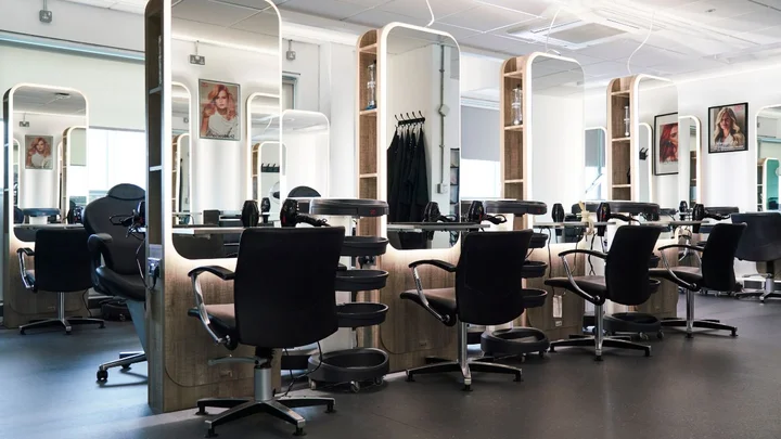 A modern hairdressing training salon with styling stations, salon chairs, mirrors, and illuminated shelving.