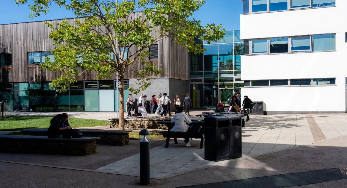 An outdoor courtyard at The Manchester College, with students walking between modern campus buildings and others sitting on benches under a tree.
