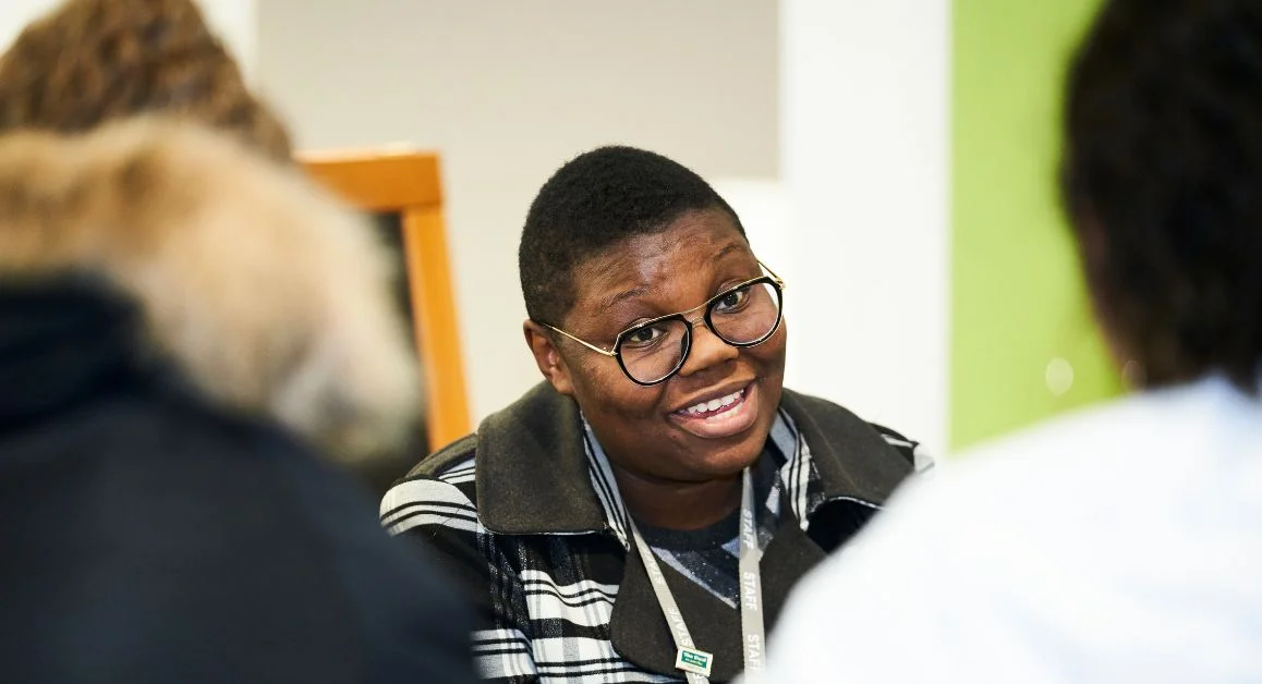 A staff member wearing a lanyard sitting at a table during a college information session, with two students seated opposite and blurred in the foreground.