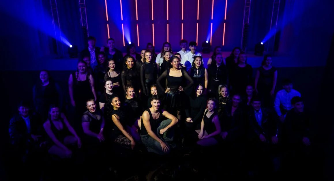 A large group of performing arts students on stage in theatrical lighting at The Manchester College, gathered in costume for a group production photo.