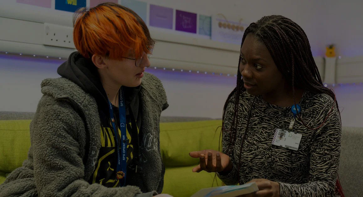 Two people sitting on a sofa at The Manchester College, engaged in conversation while looking at a document, with posters displayed on the wall behind them.