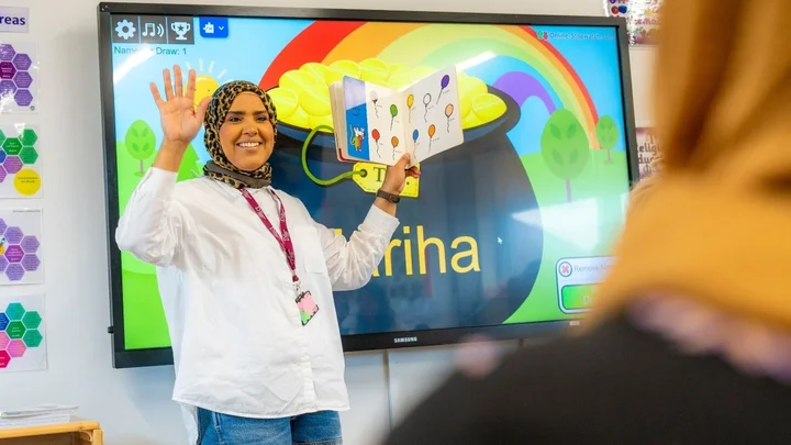 A learner in a childhood studies classroom reading a picture book aloud, standing in front of an interactive screen displaying colourful educational graphics.