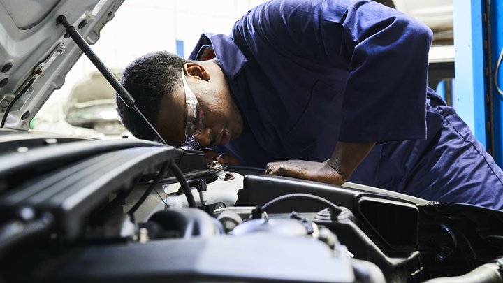 A student wearing safety goggles and overall is looking into an open car bonnet.