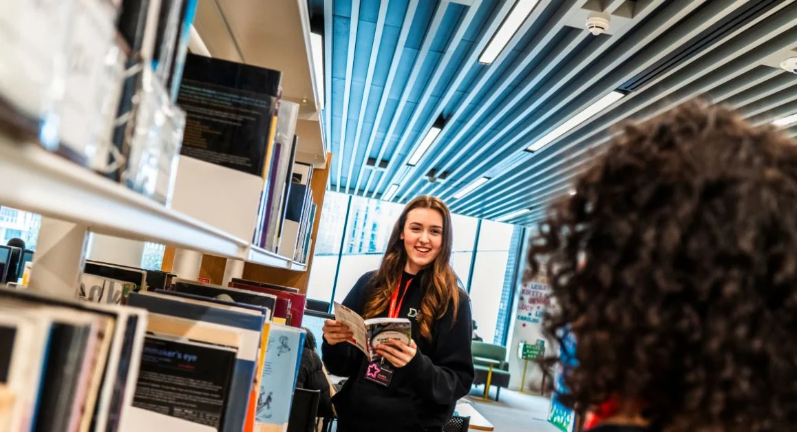 A learner from The Manchester College standing between shelves in a modern college library, opening a book while talking to another student.